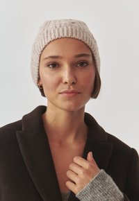 Căciulă tricotată bej cu o textură ribbată, având o potrivire strânsă. Modelul poartă o haină închisă la culoare și un pulover gri deschis. Detalii de design simple.