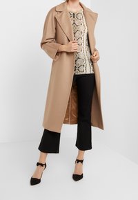 Beżowy długi płaszcz z rewersem na kołnierzu, noszony na wzorzystym topie, zestawiony z czarnymi, krótkimi spodniami i czarnymi szpilkami ze wstążkowymi ozdobami.