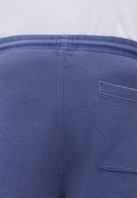 Κοντινή λήψη μπλε υφασμάτινων σορτς με ελαστική μέση και πίσω τσέπη με ραφή, φορεμένα με λευκό τοπ.