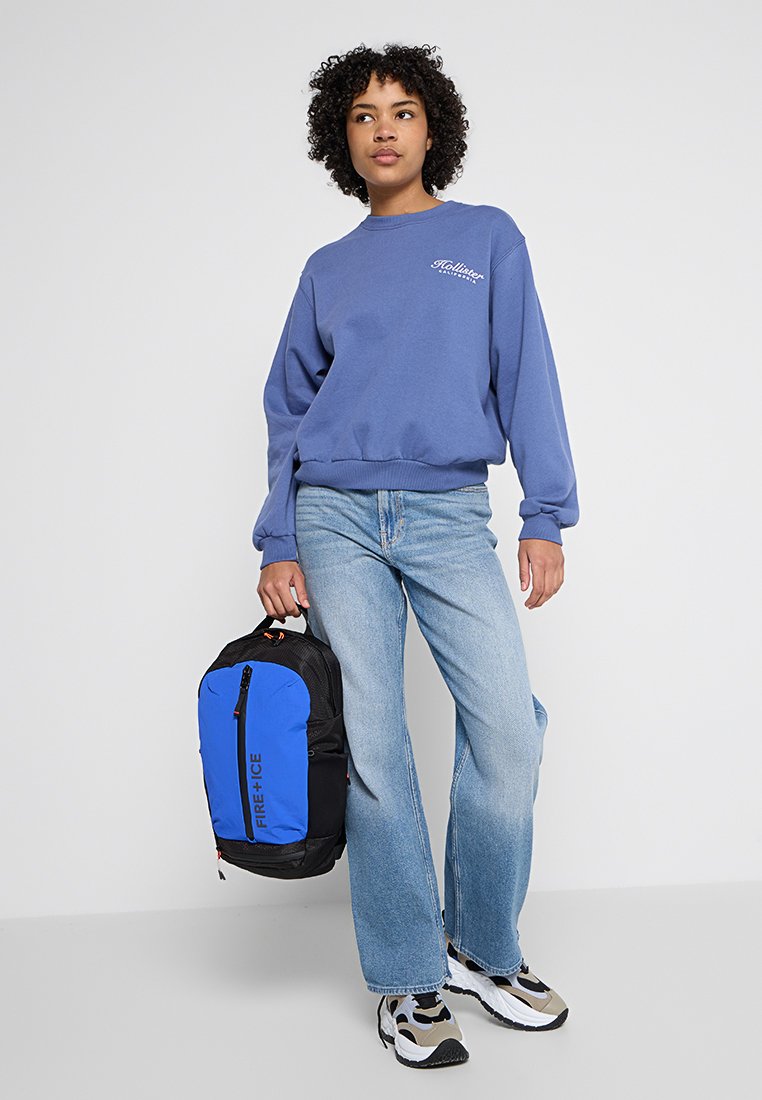 Blauer Pullover mit Logo, hellblaue Jeans und gemusterte Turnschuhe. Schwarzer Rucksack mit blauen Akzenten und einem Frontreißverschluss.