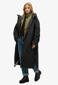 Lange zwarte buitenjas met een rits, voorzakken en een capuchon, gelaagd over een groene binnenjas en een zwarte trui, gecombineerd met blauwe jeans.