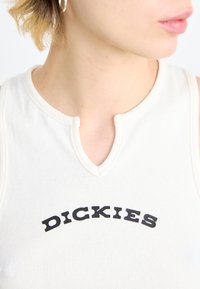 Haut sans manches, côtelé et de couleur crème avec un col en V ; présente le mot "DICKIES" en lettres noires grasses sur la poitrine.