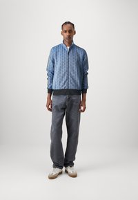 Blå mønstret zip-jakke med høj krave, ribbede manchetter og kant, parret med grå løstsiddende denimjeans og hvide sneakers.