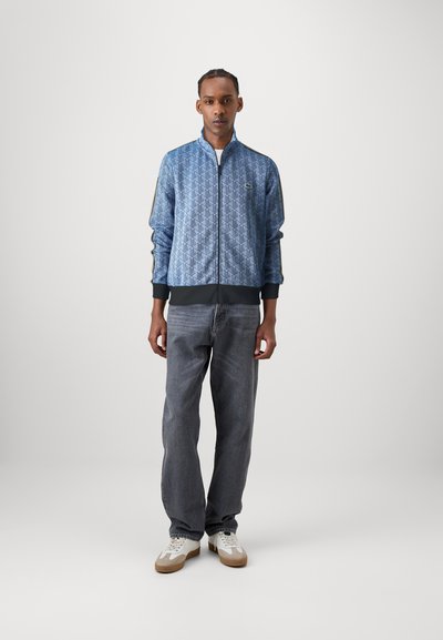 Veste zippée bleu à motifs avec un col haut, des poignets et un ourlet côtelés, associée à un jean en denim gris ample et des baskets blanches.