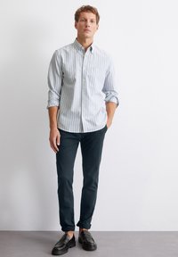 Camicia button-up a righe azzurro chiaro e bianche, maniche arrotolate, taschino sul lato sinistro del petto; abbinata a pantaloni scuri slim-fit e mocassini neri.