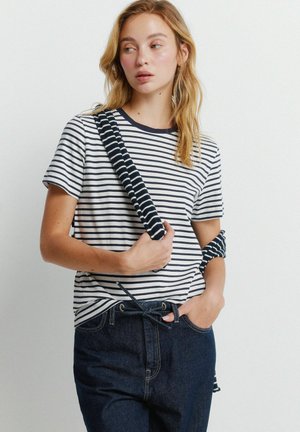 Młoda kobieta o blond włosach nosi czarno-białą pasiastą koszulkę i ciemne dżinsy, trzymając pasujący pasiasty sweter zarzucony na ramię.