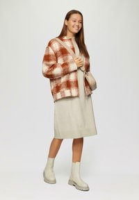 Overmaatse geruite cardigan in rust en crème, gelaagd over een lichtgrijze gebreide jurk. Afgerond met lichtbeige chunky enkellaarzen en een kleine tas.