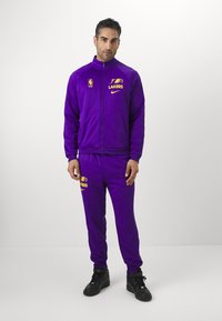 O fato de treino roxo, feito de um tecido suave, apresenta uma jaqueta com fecho éclair com o logótipo "LAKERS" e calças combinando, realçadas por sapatilhas de cano alto pretas.