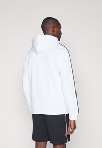 Sweatshirt alb cu glugă, realizat dintr-un amestec de bumbac, cu dungi negre pe lateral și tiv cu nervuri. Modelul poartă pantaloni scurți negri cu accente albe.