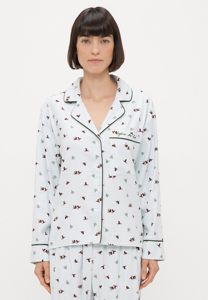 Bílé pyžamo na světle modrém pozadí, s malými červenými a zelenými vzory ptáků a stromů. Zelené lemování na límci a manžetách.