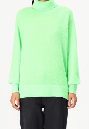 Personne portant un pull vert vif à col roulé avec des manches longues et un pantalon noir, montrée des épaules aux hanches.