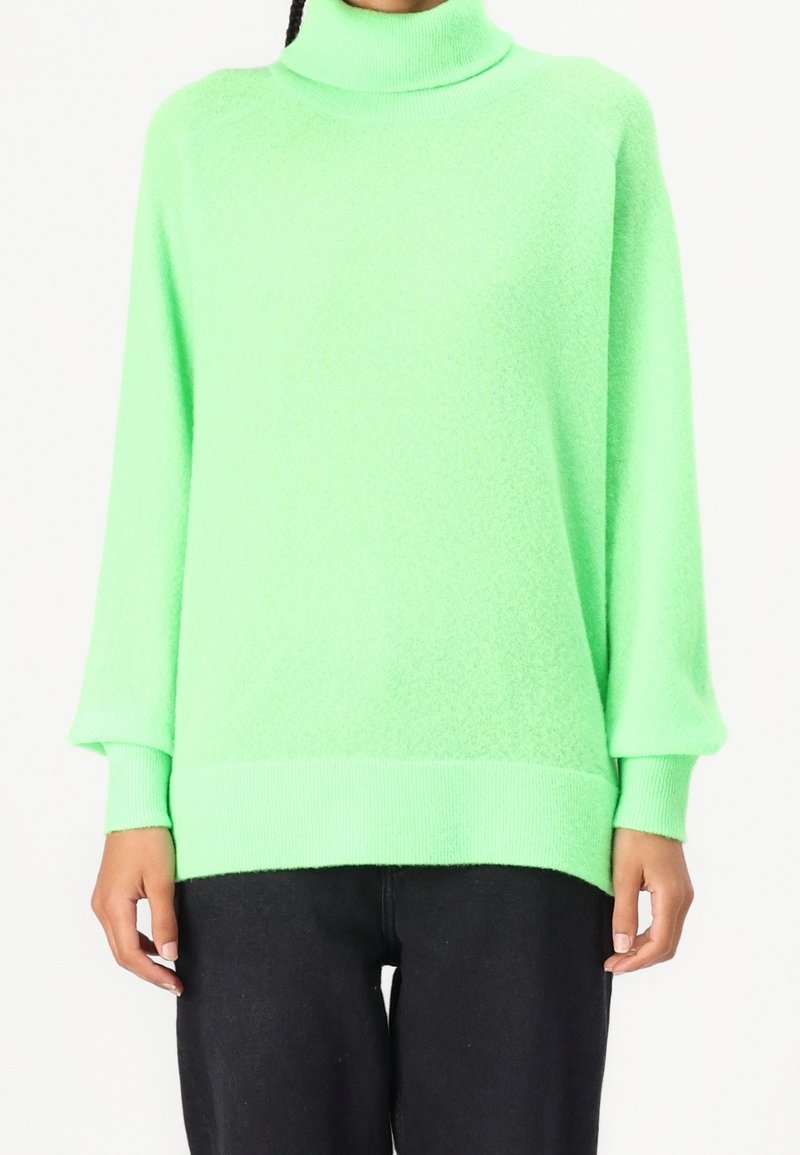 Personne portant un pull vert vif à col roulé avec des manches longues et un pantalon noir, montrée des épaules aux hanches.