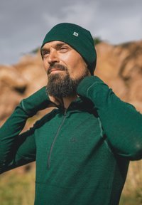 Zelený svetr na zip s vyšším límcem, dlouhými rukávy a otvory pro palce. K němu se těsně na hlavě nosí odpovídající pletená čepice.