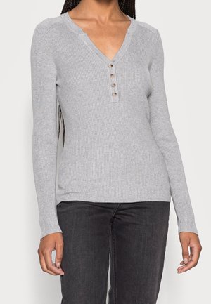 Kobieta ubrana w jasnoszary prążkowany długi rękaw henley z czterema guzikami, w połączeniu z ciemnoszarymi dżinsami.