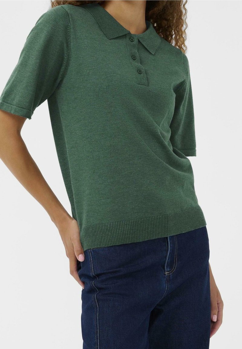 Femme portant un polo vert à manches courtes et un jean bleu foncé, une main posée sur la hanche devant un fond uni.