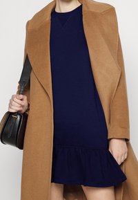 Osoba oblečená v velbloudím kabátu přes námořnické šaty s volánkovým lemem, držící černou koženou kabelku za popruh na jednom rameni.