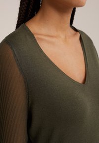 Olivgrön stickad topp med V-hals och genomskinliga, texturerade ärmar. Tyget verkar mjukt och lätt. Kanterna är snyggt avslutade.