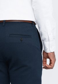 Pantaloni blu navy in tessuto strutturato, con una tasca posteriore singola con chiusura a bottone e una cintura in pelle marrone.