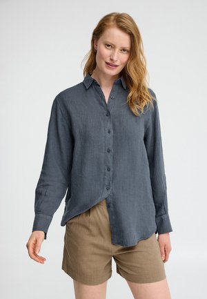 Jeune femme aux longs cheveux roux portant une chemise boutonnée gris foncé et un short marron clair, debout devant un fond blanc uni.