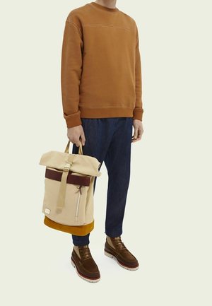 Personne portant un sweat-shirt marron, un pantalon bleu foncé et des bottes marron, tenant un sac à dos à rabat roulé beige et marron par la poignée.