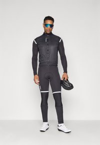 Cyklistický outfit pozostávajúci z čiernej bezrukávovej bundy a čiernych legín s bielymi reflexnými pruhmi. Doplnky zahŕňajú prilbu a slnečné okuliare.