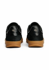 Paar schwarze Sneaker mit niedrigem Schaft und braunen Sohlen, Rückansicht, mit kleinem goldfarbenem Logo auf den Fersenlaschen vor weißem Hintergrund.