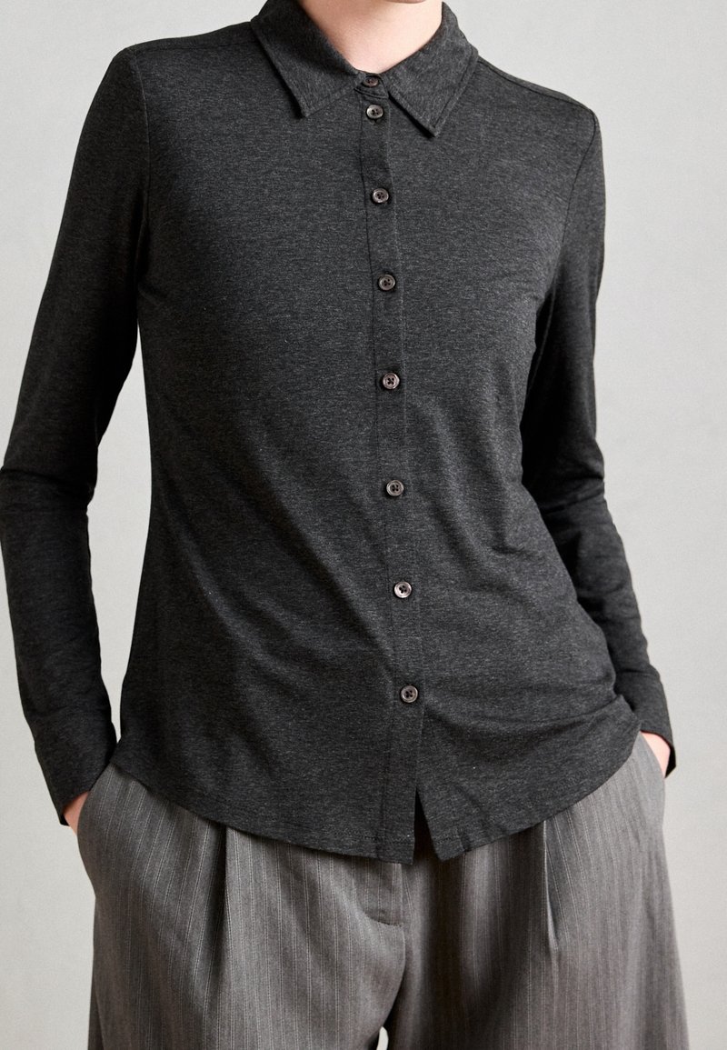 Personne portant une chemise à manches longues gris foncé boutonnée avec col et un pantalon gris à fines rayures, mains dans les poches, debout devant un fond uni.