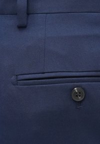 Tamsiai mėlyni kelnės su lygiu audiniu turi šoninę kišenę ir juodą keturiais skylutėmis mygtuką. Audinys pasižymi subtiliu įstrižiniu pynimu.