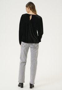 Schwarzes Samtoberteil mit langen Ärmeln, einem Rückenschlitz-Detail und gerafften Schulternähten, kombiniert mit hellgrauen, geraden Jeans und Stiefeln mit Absatz.