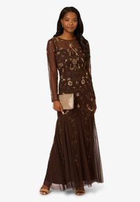Hnědé večerní šaty s dlouhými rukávy, průhledným přehozem a zlatou květinovou výšivkou. Modelka drží zlatou kabelku a má na sobě sandály na otevřenou špičku.