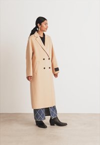 Donna in piedi che indossa un lungo cappotto beige doppiopetto, jeans a gamba larga con fantasia, stivali neri e orecchini dorati, contro uno sfondo bianco.