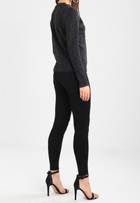 Schwarzer Strickpullover mit Rundhalsausschnitt, taillierter Form und geripptem Saum. Kombiniert mit schwarzen Skinny-Jeans und schwarzen High-Heels.