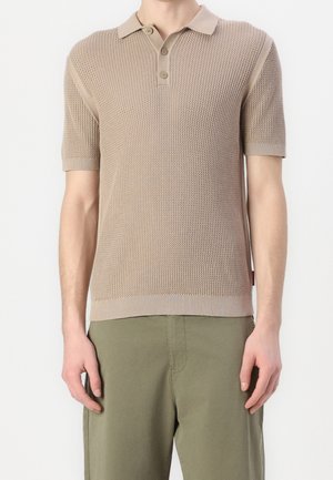 Homme portant un polo en maille à manches courtes beige texturé avec trois boutons et un pantalon vert olive, debout devant un fond uni.