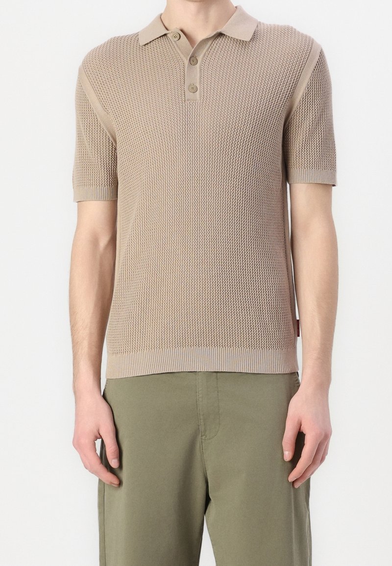 Homme portant un polo en maille à manches courtes beige texturé avec trois boutons et un pantalon vert olive, debout devant un fond uni.