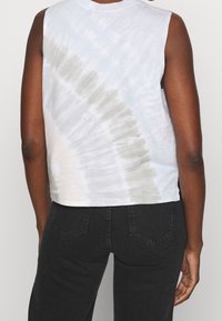 Zkrácený bezrukávový tričko ve světle modro-šedém tie-dye vzoru, vyrobené z měkkého materiálu, spárované s tmavými džíny.