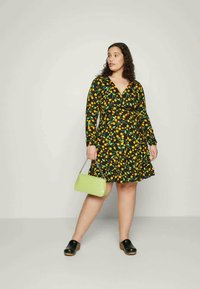 Veste preta em envoltório com padrão de frutas amarelas, mangas longas e saia ampla; combinada com uma pequena mala verde e tamancos pretos.