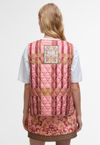 Colete acolchoado em tons de rosa, com um padrão de diamantes, adornado com um colorido patch de flores nas costas. Usado sobre uma t-shirt branca e uma saia floral.