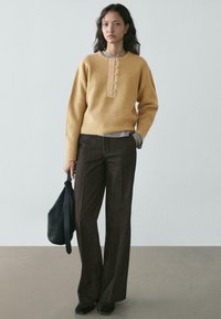 Maglione a coste color senape con una piega centrale con bottoni, abbinato a pantaloni marroni su misura e una borsa nera testurizzata.