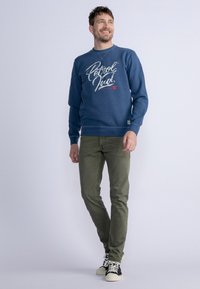 Sudadera azul con gráfico en blanco y rojo que dice "Petrol", combinada con pantalones verde oliva y zapatillas en blanco y negro. Estilo casual, corte relajado.