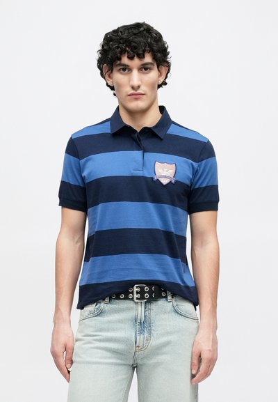 Μπλε ναυτικό και ανοικτό μπλε ριγωτό πόλο πουκάμισο με γιακά, κοντά μανίκια και λογότυπο στο αριστερό στήθος. Φοράται με ανοιχτόχρωμο τζιν και ζώνη.