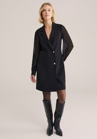 Rochie neagră cu dublu buton, cu decupaje la umeri, având mâneci lungi transparente și nasturi aurii, asortată cu cizme negre lucioase și înalte.