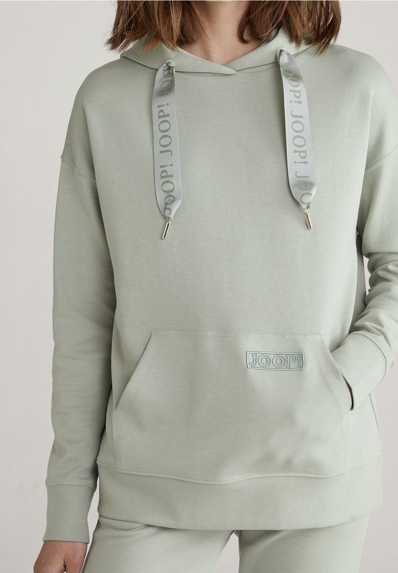 Femme portant un sweat à capuche vert sauge clair avec poche avant et cordons assortis estampillés "JOOP!".