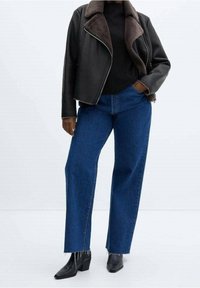 Veste en simili cuir noir avec col en shearling marron, associée à un jean large bleu et des bottines noires à talons.