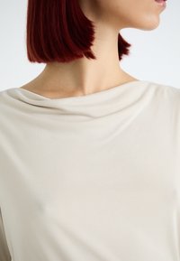 Beige drapertopp med myk, jevn tekstur og båthals, som fremhever kragebeinet og skulderen med minimal detalj.