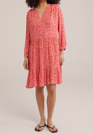 Roze jurk met bladerenpatroon, v-hals en lange mouwen. Vloeiende stof, gelaagde rok en aansluitend in de taille. Gdragen met zwarte slippertjes.