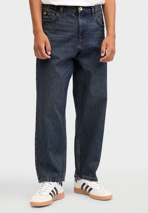 Homme portant un jean droit bleu foncé et des baskets blanches avec des rayures noires, debout sur un fond clair.