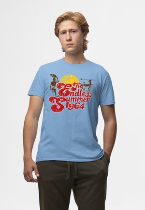 Ung mand med blondt hår iført en lyseblå T-shirt med rød tekst "The Endless Summer 1964" og en gul solgrafik.