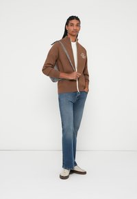 Brun zip-up genser med høy krage, parret med mørkeblå jeans og kremfargede sneakers. Genseren har en logo detalj på brystet.