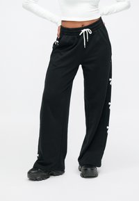 Black sweatpants made of soft fabric, featuring a drawstring waist and side accents of white bows along the legs. Relaxed fit, wide leg.