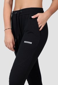 Čierne bežecké nohavice vyrobené z mäkkej látky s elastickým pásom na šnúrku, bočnými vreckami a malou logovou nášivkou pri ľavom bedre.